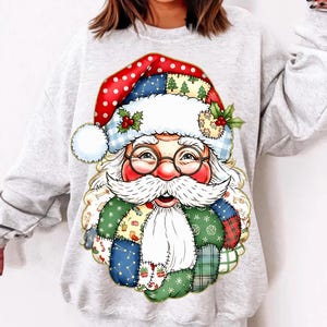 Pode incluir: Um moletom cinza claro com um gráfico colorido do Papai Noel. O design do Papai Noel inclui um chapéu vermelho e branco com bolinhas, óculos e uma longa barba branca. O rosto do Papai Noel é cercado por um design de patchwork de várias cores e padrões.
