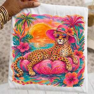 Pode incluir: Camiseta branca com uma ilustração colorida de um leopardo usando óculos de sol e um chapéu rosa, relaxando em um flutuador rosa no oceano. O fundo inclui um pôr do sol, palmeiras e flores tropicais.