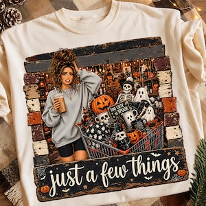 May include: A cream-colored t-shirt featuring a graphic of a person holding a coffee cup next to a shopping cart filled with Halloween items like pumpkins, skulls, and ghosts. The text reads "Too cute to spook" and "just a few things."