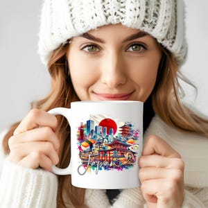 May include: White ceramic mug featuring a colorful watercolor design of Japanese landmarks, including a red sun, skyscrapers, and temples. The word "Japan" is written in a script font. The mug is held by a person wearing a white knit hat.