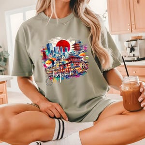 May include: Sage green t-shirt featuring a colorful graphic of Japanese landmarks, including a red sun, pagodas, and city buildings. The word "Japan" is written in a cursive font across the graphic. The shirt is a casual, comfortable style.