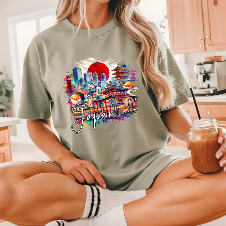 May include: Sage green t-shirt featuring a colorful graphic of Japanese landmarks, including a red sun, pagodas, and city buildings. The word "Japan" is written in a cursive font across the graphic. The shirt is a casual, comfortable style.