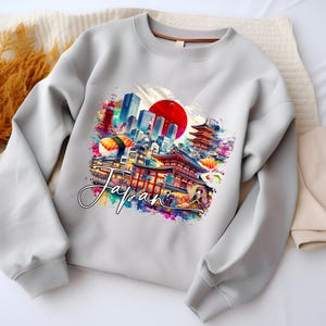 May include: A light gray sweatshirt featuring a colorful graphic of Japanese landmarks, including a red sun, pagodas, and cityscapes. The word "Japan" is written in a cursive font across the design. The sweatshirt has long sleeves and a crew neck.