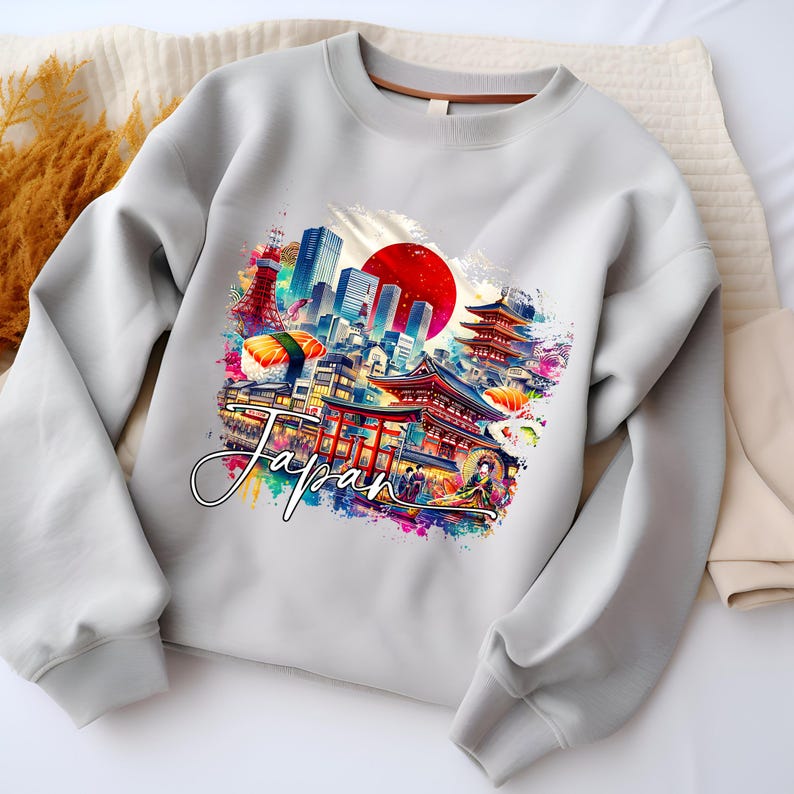 May include: A light gray sweatshirt featuring a colorful graphic of Japanese landmarks, including a red sun, pagodas, and cityscapes. The word "Japan" is written in a cursive font across the design. The sweatshirt has long sleeves and a crew neck.