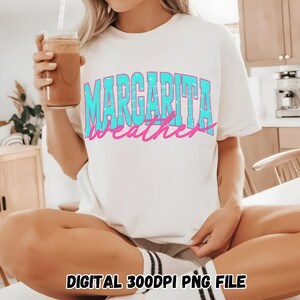 May include: White t-shirt with the text "MARGARITA weather" in turquoise and pink. The shirt is a relaxed fit. A person is holding a glass of a brown beverage with a straw.