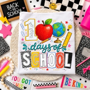 May include: A white t-shirt featuring a colorful graphic with the text 100 days of SCHOOL. The design includes a large red apple, a globe on a stand, a yellow ruler, a pencil, a lightbulb, and the number 1 with 123 written inside it.