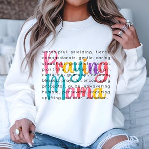 Peut inclure: Sweat-shirt blanc avec le mot "Priant" &eacute;crit en couleurs arc-en-ciel. Le mot est entour&eacute; d'une liste d'adjectifs positifs d&eacute;crivant une personne qui prie.