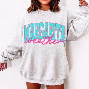 May include: A light gray sweatshirt with the words "MARGARITA weather" printed across the front in turquoise and pink. The sweatshirt has a crew neck and long sleeves.