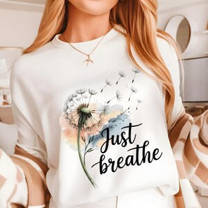 K&ouml;nnte beinhalten: Wei&szlig;es Sweatshirt mit einem L&ouml;wenzahnmuster und dem Text "Just breathe" in schwarzer Schrift.