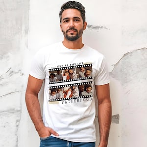 Pu&ograve; includere: T-shirt bianco con un design a striscia di pellicola con foto di famiglia e la scritta "AIN'T NO HOOD LIKE FATHERHOOD". La maglietta &egrave; indossata da una persona con capelli scuri e barba.