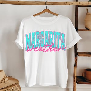 May include: White t-shirt with the words "MARGARITA weather" in a turquoise and pink distressed font. The shirt is hanging on a wooden hanger against a neutral background.