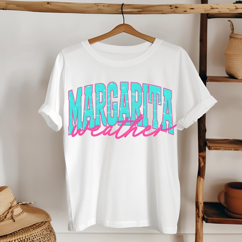 May include: White t-shirt with the words "MARGARITA weather" in a turquoise and pink distressed font. The shirt is hanging on a wooden hanger against a neutral background.