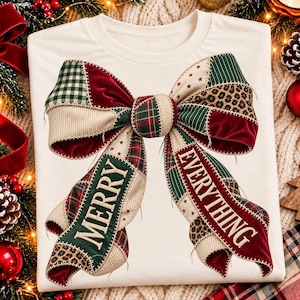 May include: A cream-colored t-shirt featuring a large, central graphic of a patchwork-style bow. The bow is composed of various fabric patterns including green plaid, red velvet, and leopard print, with the text MERRY EVERYTHING printed across it.