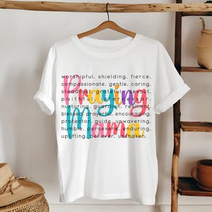 Peut inclure: T-shirt blanc avec un motif graphique color&eacute; qui dit "Praying Mama" et une liste de mots d&eacute;crivant les qualit&eacute;s d'une m&egrave;re qui prie. Les mots sont de diff&eacute;rentes couleurs, notamment rose, bleu, vert et jaune.