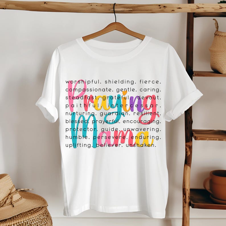 Peut inclure: T-shirt blanc avec un motif graphique color&eacute; qui dit "Praying Mama" et une liste de mots d&eacute;crivant les qualit&eacute;s d'une m&egrave;re qui prie. Les mots sont de diff&eacute;rentes couleurs, notamment rose, bleu, vert et jaune.
