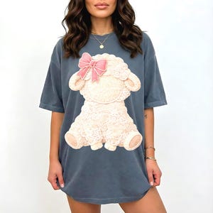 Pode incluir: Camiseta azul ardósia com um grande gráfico de um cordeiro com um laço rosa. O cordeiro tem um corpo creme com detalhes de renda. A camiseta tem mangas curtas e um corte folgado.