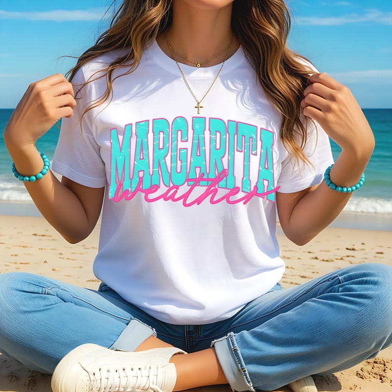 May include: White t-shirt with the words "MARGARITA weather" in turquoise and pink. The person is wearing blue jeans, white sneakers, and turquoise bracelets. The background shows a beach and ocean.