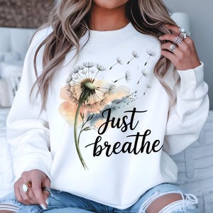 K&ouml;nnte beinhalten: Wei&szlig;er Pullover mit einem Aquarell-L&ouml;wenzahnmuster und dem Text "Just breathe".