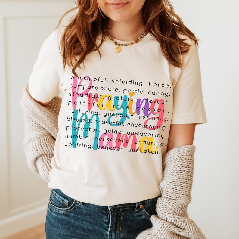 Peut inclure: Un t-shirt blanc avec le mot "Praying" &eacute;crit en lettres color&eacute;es. Le mot est d&eacute;fini avec des attributs positifs tels que "worshipful", "fierce", "compassionate", "steadfast", "grateful", "devout", "intercessor", "nurturing", "guardian", "resilient", "blessed", "prayerful", "encouraging", "protector", "guide", "unwavering", "humble", "persevere", "enduring", "uplifting", "believer", et "unshaken".