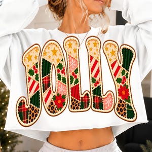 Pode incluir: Uma camisola branca de manga comprida e corte curto com a palavra JOLLY em letras grandes estilo patchwork. As letras estão preenchidas com vários padrões, incluindo estrelas douradas, riscas vermelhas e brancas, xadrez verde e bagas de azevinho.