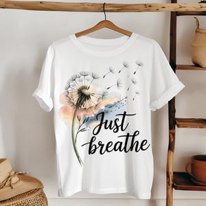 K&ouml;nnte beinhalten: Wei&szlig;es T-Shirt mit einem Aquarell-L&ouml;wenzahnmuster und dem Text "Just breathe" in Schwarz.