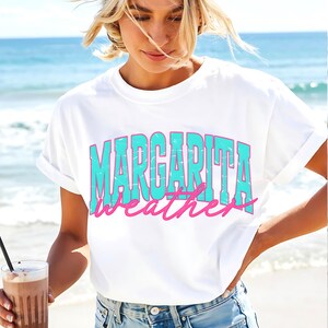 May include: White t-shirt with the words "MARGARITA weather" in turquoise and pink lettering. The shirt has rolled-up sleeves and is worn by a person on a beach. A drink with a straw is held in the person's hand.