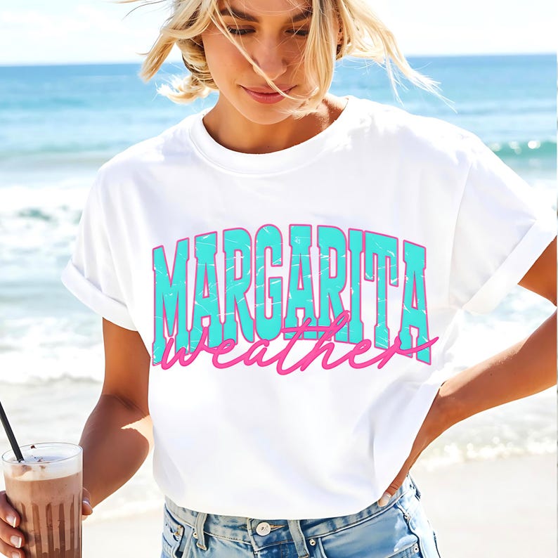 May include: White t-shirt with the words "MARGARITA weather" in turquoise and pink lettering. The shirt has rolled-up sleeves and is worn by a person on a beach. A drink with a straw is held in the person's hand.