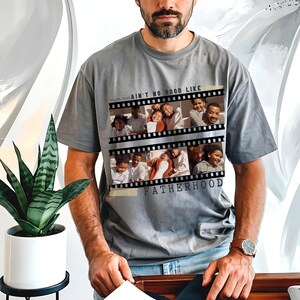 Pu&ograve; includere: T-shirt grigio con un design a striscia di pellicola con foto di famiglia e la scritta "AIN'T NO HOOD LIKE FATHERHOOD". La maglietta ha una vestibilit&agrave; ampia e maniche corte.
