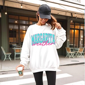 May include: White sweatshirt with the text "Margarita Weather" in turquoise and pink. The person in the image is wearing a black baseball cap and black leggings. A coffee cup is held in the person's hand.
