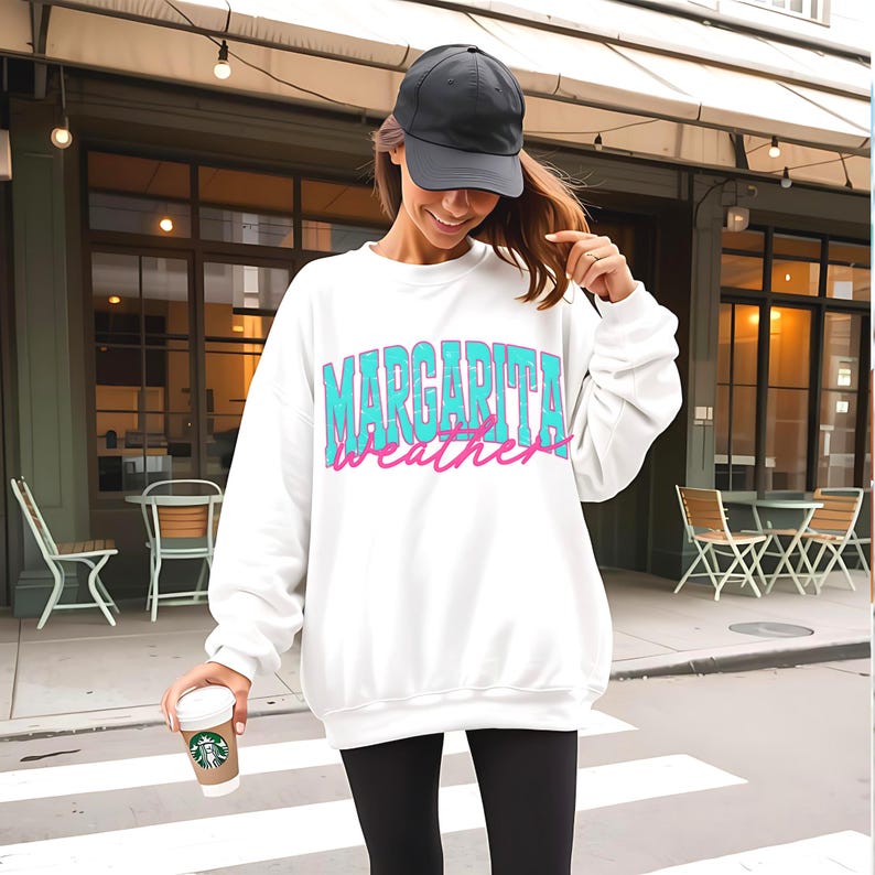 May include: White sweatshirt with the text "Margarita Weather" in turquoise and pink. The person in the image is wearing a black baseball cap and black leggings. A coffee cup is held in the person's hand.