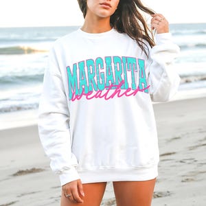 May include: White sweatshirt with the words "MARGARITA WEATHER" in turquoise and pink lettering. The sweatshirt is a crew neck style and is being worn on a beach.