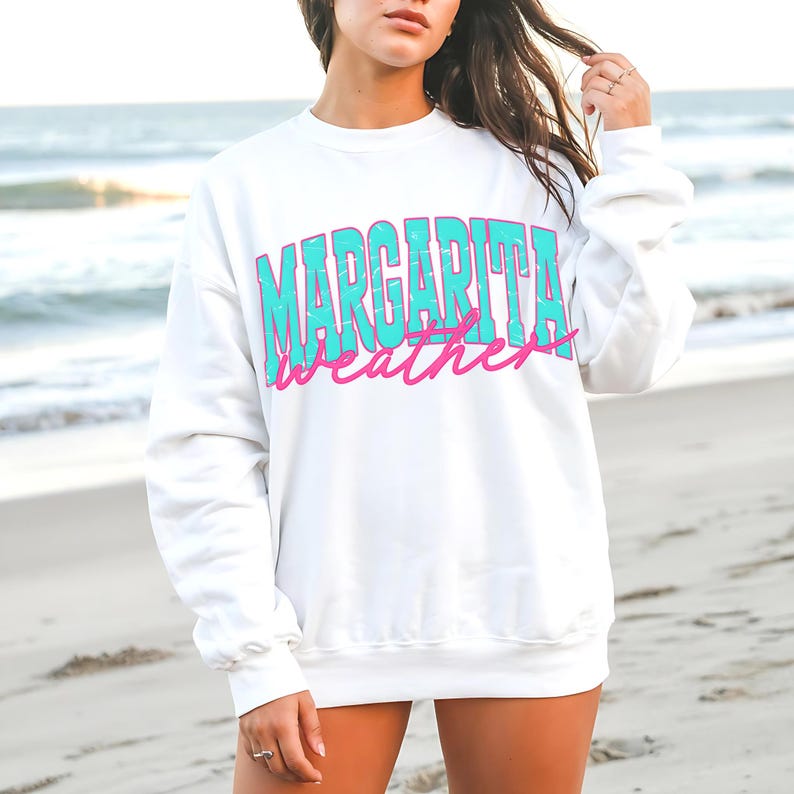 May include: White sweatshirt with the words "MARGARITA WEATHER" in turquoise and pink lettering. The sweatshirt is a crew neck style and is being worn on a beach.