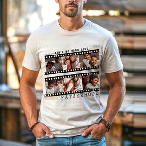 Pu&ograve; includere: Una maglietta beige chiaro con un design a striscia di pellicola con foto di famiglia. Il testo "AIN'T NO HOOD LIKE FATHERHOOD" &egrave; stampato sulla maglietta. La maglietta &egrave; a maniche corte e ha uno scollo tondo.
