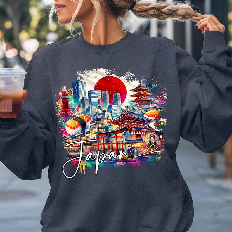 May include: Dark gray sweatshirt featuring a colorful graphic of a Japanese cityscape with a red sun, pagodas, and the word "Japan" in a cursive font. The design includes sushi and a geisha. The sweatshirt has long sleeves and a crew neck.