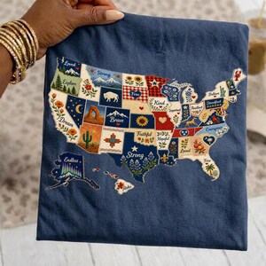 May include: A navy blue decorative pillow featuring an embroidered map of the United States. Each state is decorated with floral and nature-themed designs and words like "Dream", "Strong", and "Free".