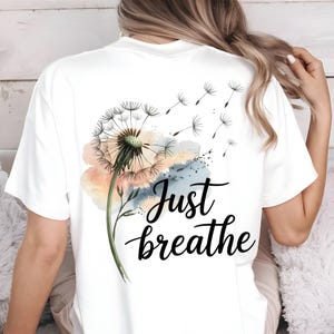 K&ouml;nnte beinhalten: Wei&szlig;es T-Shirt mit einem Aquarell-L&ouml;wenzahnmuster und dem Text "Just breathe" auf der R&uuml;ckseite.