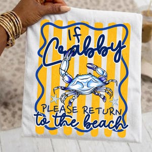 Puede incluir: Camiseta blanca con una ilustración de cangrejo azul y blanco y el texto "If Crabby Please Return to the Beach". El diseño incluye un fondo a rayas amarillas y azules y letras azules. La camiseta está sujeta por una persona.
