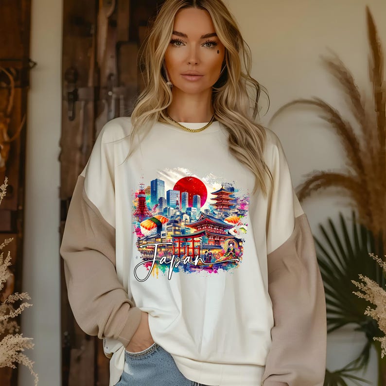 May include: Cream-colored sweatshirt with tan sleeves featuring a colorful graphic of Japanese landmarks, sushi, and a rising sun. The word "Japan" is written in cursive below the graphic. The sweatshirt is worn by a person.