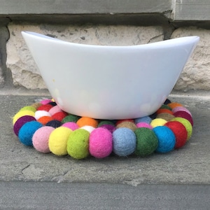 May include: A white ceramic bowl sits on a colorful felt coaster. The coaster is made of a circle of felt balls in various colors, including pink, blue, green, yellow, and red.