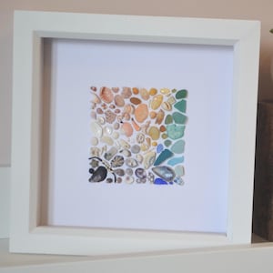 May include: A white framed artwork featuring a square arrangement of seashells in various colors. A small succulent plant in a white pot sits on a wooden block next to the artwork. The artwork is on a white shelf.