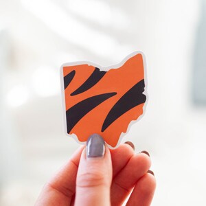 Op de afbeelding: Een sticker in de vorm van de staat Ohio, met een oranje achtergrond en zwarte strepen. De sticker wordt vastgehouden door de hand van een persoon.