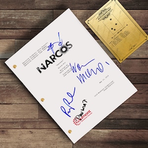 May include: A white script for the TV show "Narcos" featuring multiple blue ink signatures. The script displays the show's title and episode details. A gold-coloured "Autograph Key" card is in the upper right. The background is a wooden surface.