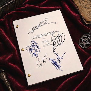 Könnte beinhalten: Ein weißes Drehbuch mit dem Titel "Supernatural" mit mehreren Unterschriften in blauer und schwarzer Tinte. Das Drehbuch wird mit goldenen Reißnägeln zusammengehalten und liegt auf einer tiefroten Samtoberfläche. Ein Pentagramm-Anhänger ist im Hintergrund zu sehen.