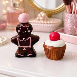 Pode incluir: Dois itens decorativos: um homem de gengibre com uma bola rosa em cima e um cupcake com uma bola vermelha em cima. O homem de gengibre é castanho com detalhes rosa. O cupcake tem cobertura branca e uma base castanha.