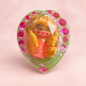 May include: A heart-shaped ring featuring a central clear faceted dome that encases a small doll with bright yellow hair and a pink outfit. The outer edge of the heart is decorated with multiple round, vibrant pink rhinestones set in green.
