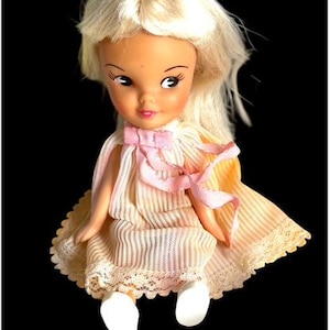 May include: A vintage doll with blonde hair and a pink bow. The doll wears a cream-colored dress with a striped pattern and lace trim. The doll's shoes are white.