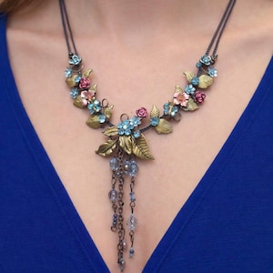 Puede incluir: Un collar floral con acabado bronce. El collar presenta flores y hojas rosas, azules y doradas. Cuentas azules colgantes cuelgan del centro del collar. El collar se usa sobre un fondo azul.