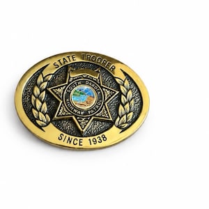 以下が含まれることがあります： 中央に6つの突起がある星のエンブレムと「South Dakota Highway Patrol」の文字が刻まれた楕円形の真鍮製ベルトバックル。上部には「State Trooper」、下部には「Since 1938」の文字が配置され、月桂樹の葉の装飾が施されています。