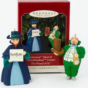 Puede incluir: Un juego de dos adornos navideños con el tema "El Mago de Oz". Un adorno presenta una figura con un atuendo azul y verde que sostiene un certificado. El otro adorno presenta una figura verde con un reloj de bolsillo y un sombrero de copa marrón. La caja dice "Keepsake Ornament".