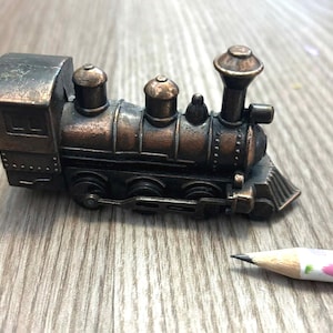 May include: A detailed, antique-style train pencil sharpener in shades of bronze and brown. The miniature train features intricate detailing, including a smokestack, wheels, and a cabin. A sharpened pencil with a pink and green floral design is next to the train.